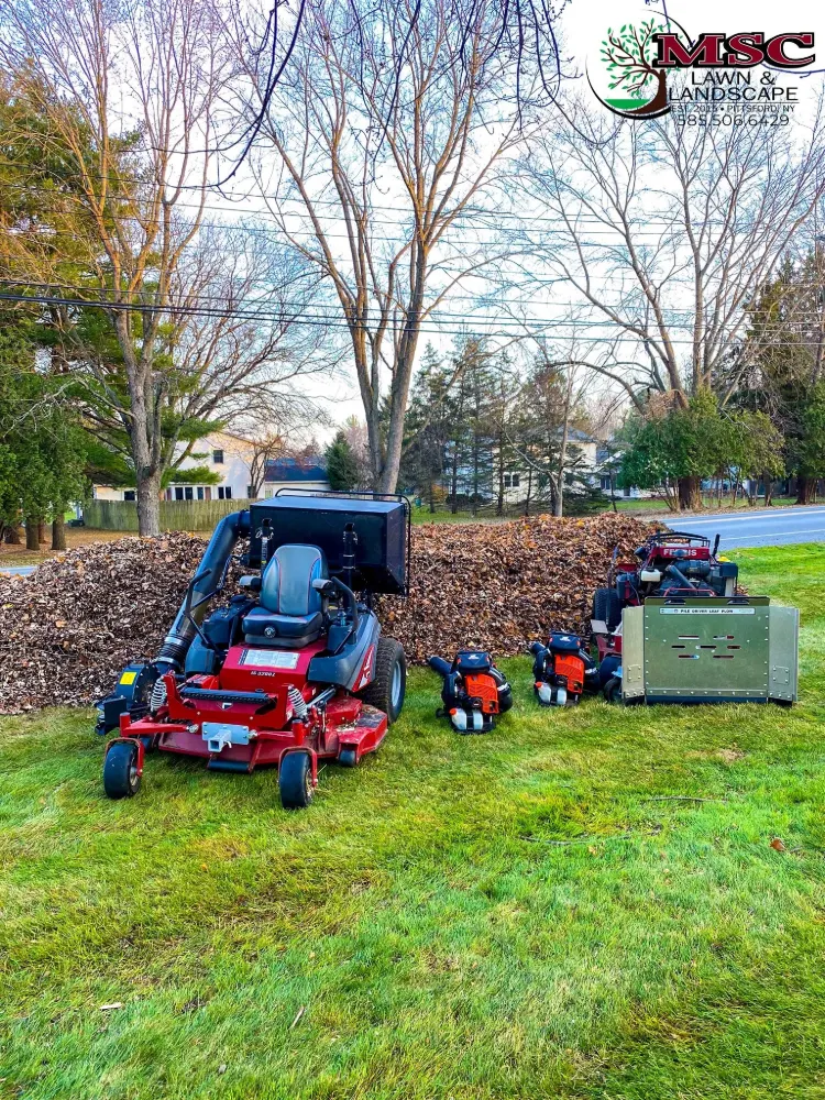 A pile of fall leaves beside a zero-turn mower with a leaf vacuum, two backpack blowers and a stand-on mower with a leaf plow